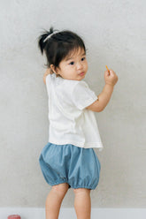 Baby girl wearing Avauma Oat Latte short-sleeve cardigan with blue bloomers, walking indoors.